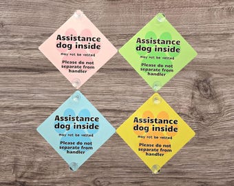 May include: Four diamond-shaped signs in pink, green, blue, and yellow, each with the text "Assistance dog inside." The signs also include the text "may not be vested" and "Please do not separate from handler."