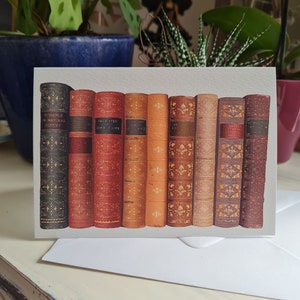 May include: A greeting card featuring a vintage illustration of a row of ten books with gold lettering on their spines. The books are arranged in a bookshelf style, with the spines facing the viewer. The card is a light grey colour with a subtle texture.