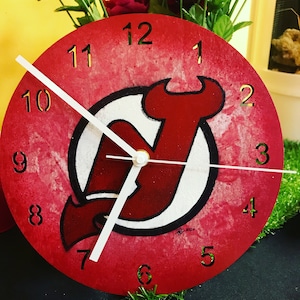 May include: Red and white clock with a New Jersey Devils logo. The clock has black numbers and a white background. The logo is a red 'N' with a white outline. The clock is 12 inches in diameter.