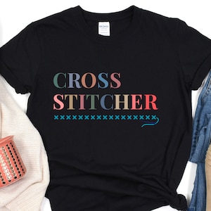 May include: Black t-shirt with the text "CROSS STITCHER" in rainbow colours. The text is followed by a row of blue "x"s and a blue needle and thread design.