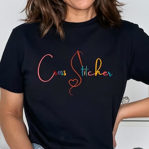 May include: Black t-shirt with a colorful graphic that says "Cross Stitcher" with a needle and thread design and a heart.