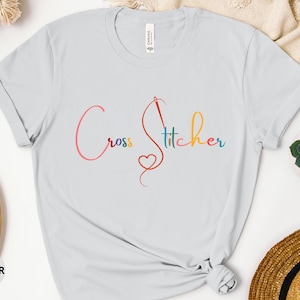 Può includere: Una maglietta blu chiaro con il testo "Cross Stitcher" in lettere corsive colorate. Un design stilizzato di ago e filo è incorporato nel testo.
