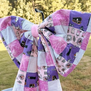 May include: A large fabric bow featuring a patchwork design with purple, pink, and white colours. The pattern includes illustrations of ghosts, coffins, tombstones, and spider webs. The bow is constructed from quilted fabric squares and has a solid pink centre.