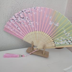 Op de afbeelding: Decoratieve handwaaier in roze, groene en gele tinten. De waaier is versierd met een bloemmotief van kersenbloesems en vlinders. Hij heeft een houten handvat en een roze kwast.