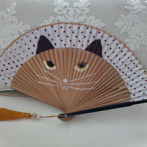 Peut inclure: Éventail décoratif avec un motif de visage de chat. Le ventilateur présente un fond beige avec un visage de chat peint dessus, comprenant des oreilles marron et des yeux jaunes. La bordure du ventilateur est blanche avec des pois noirs. Une gland jaune est suspendu au ventilateur.