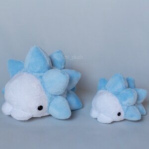 Puede incluir: Dos peluches con forma de criatura azul y blanca con pinchos. El peluche m&aacute;s grande est&aacute; tumbado y el m&aacute;s peque&ntilde;o est&aacute; de pie. Los peluches est&aacute;n hechos de tela suave y peluda y tienen ojos negros.