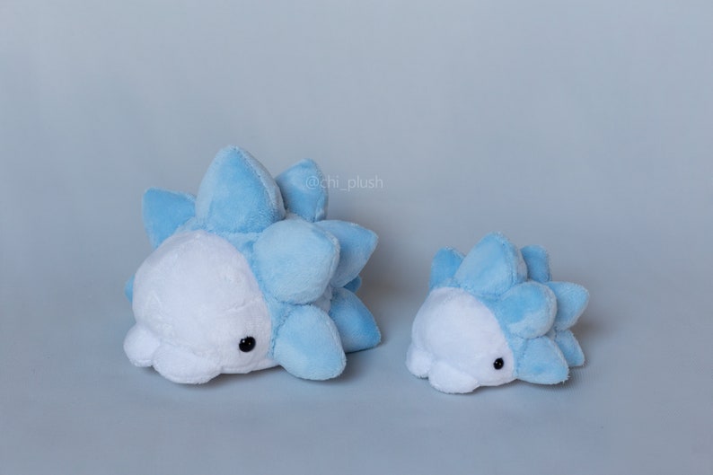Puede incluir: Dos peluches con forma de criatura azul y blanca con pinchos. El peluche m&aacute;s grande est&aacute; tumbado y el m&aacute;s peque&ntilde;o est&aacute; de pie. Los peluches est&aacute;n hechos de tela suave y peluda y tienen ojos negros.