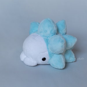 Puede incluir: Un juguete de peluche azul claro y blanco con forma de criatura redonda y espinosa. El juguete tiene una cara blanca con ojos negros y un cuerpo azul claro con protuberancias redondeadas y espinosas.