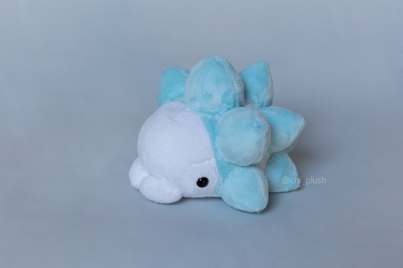 Puede incluir: Un juguete de peluche azul claro y blanco con forma de criatura redonda y espinosa. El juguete tiene una cara blanca con ojos negros y un cuerpo azul claro con protuberancias redondeadas y espinosas.