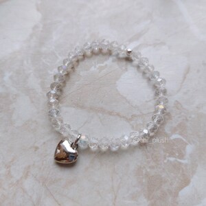 May include: A clear crystal beaded bracelet with a silver heart charm. The bracelet is on a white marble surface.