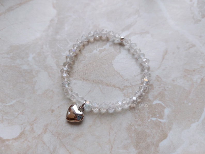 May include: A clear crystal beaded bracelet with a silver heart charm. The bracelet is on a white marble surface.