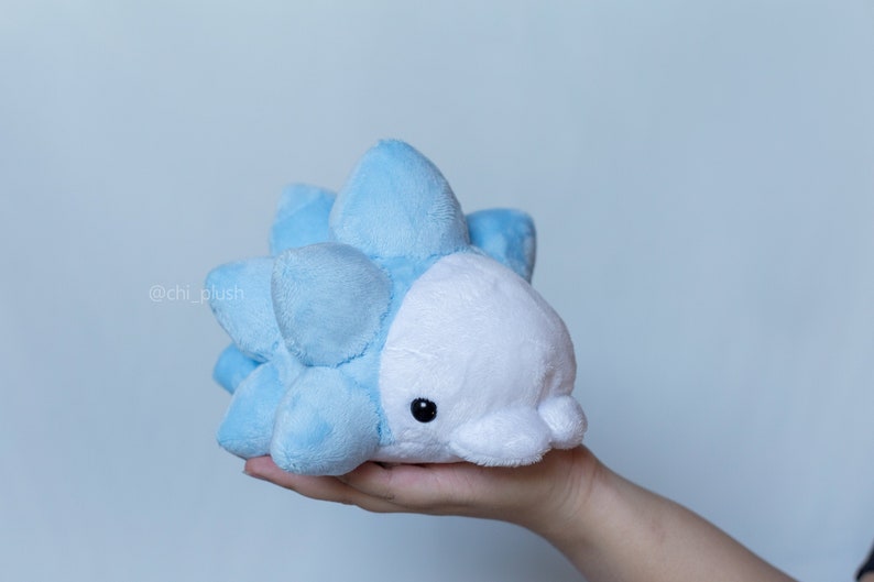 Puede incluir: Un juguete de peluche azul claro y blanco que se asemeja a una criatura marina espinosa. El juguete tiene una cara blanca con ojos negros y una peque&ntilde;a boca.