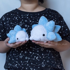 Puede incluir: Dos peluches azules y blancos con forma de criatura redonda y espinosa. Los peluches est&aacute;n siendo sostenidos por una persona que lleva una camiseta negra con un patr&oacute;n de estrellas blancas. El texto "@chi_plush" es visible en la esquina inferior derecha de la imagen.