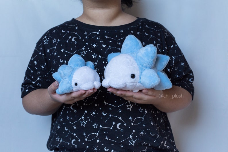 Puede incluir: Dos peluches azules y blancos con forma de criatura redonda y espinosa. Los peluches est&aacute;n siendo sostenidos por una persona que lleva una camiseta negra con un patr&oacute;n de estrellas blancas. El texto "@chi_plush" es visible en la esquina inferior derecha de la imagen.