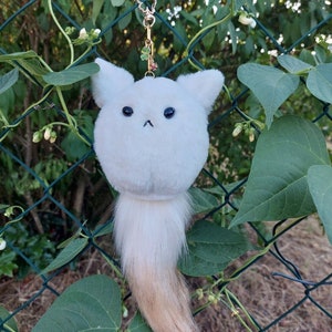 Puede incluir: Un llavero blanco y esponjoso con una cola larga, blanca y marrón. El llavero está colgado de una cadena dorada con un dije de trébol.