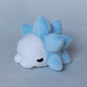 Puede incluir: Un juguete de peluche azul y blanco con forma de criatura redonda y espinosa. El juguete tiene una cara blanca con ojos negros y una peque&ntilde;a boca.