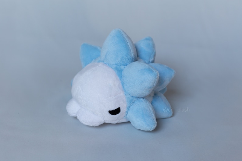 Puede incluir: Un juguete de peluche azul y blanco con forma de criatura redonda y espinosa. El juguete tiene una cara blanca con ojos negros y una peque&ntilde;a boca.