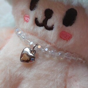 May include: A white plush toy with a heart-shaped charm necklace. The necklace is made of clear beads and a silver heart pendant.