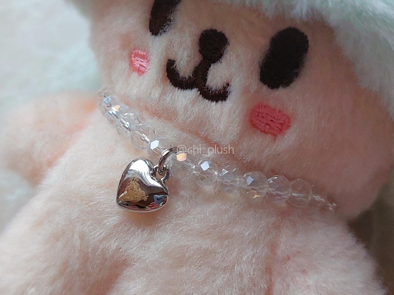 May include: A white plush toy with a heart-shaped charm necklace. The necklace is made of clear beads and a silver heart pendant.