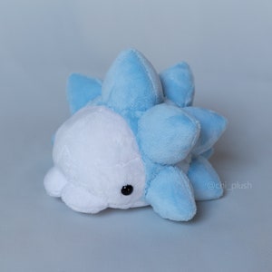 Puede incluir: Un juguete de peluche azul y blanco con una espalda puntiaguda. El juguete tiene un solo ojo negro y una peque&ntilde;a boca.