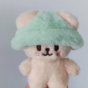 May include: A small, plush, white bear wearing a light green fuzzy hat. The bear has embroidered eyes and a small, pink nose.
