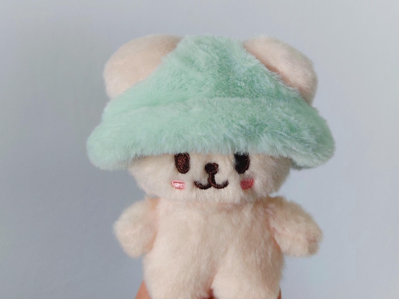 May include: A small, plush, white bear wearing a light green fuzzy hat. The bear has embroidered eyes and a small, pink nose.