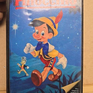 Könnte beinhalten: Ein Vintage-VHS-Kassetten-Etui mit Walt Disneys Pinocchio. Das Etui ist schwarz mit blauem Hintergrund und einem bunten Bild von Pinocchio, der mit Jiminy Cricket geht. Der Text "The Original Animated Classic!" ist in der unteren rechten Ecke des Etuis aufgedruckt.