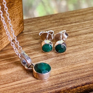 May include: A silver necklace with a round, emerald-green pendant and matching stud earrings. The jewellery set is displayed on a wooden surface, showcasing the vibrant green gemstones and the reflective silver metal.