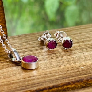 May include: A set of silver earrings and a necklace featuring a pink gemstone. The earrings are stud earrings with a round pink gemstone. The necklace has a pendant with a round pink gemstone.