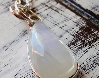 Puede incluir: Un collar de cadena de plata con un colgante de piedra de luna blanca. El colgante tiene forma ovalada y una superficie lisa y pulida.