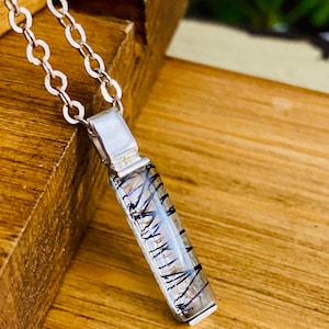 Könnte beinhalten: Eine silberne Kettenkette mit einem Anhänger aus klarem Quarzkristall mit schwarzen Einschlüssen. Der Anhänger ist rechteckig und hat eine silberne Öse.