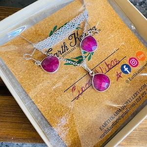 Könnte beinhalten: Ein silbernes Halsketten- und Ohrring-Set mit rosa Edelstein-Anhängern. Der Schmuck ist in einer weißen Schachtel mit einer braunen Karte verpackt, auf der "Earth Karma Jewellery and Boutique" und "Earthkarmajewellery.com" steht.