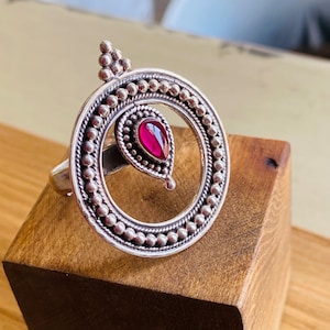 May include: A silver ring with a circular design and a pink gemstone in the center. The ring is set on a wooden surface.