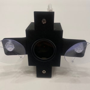 May include: A black camera with a lens and two suction cups attached to the sides. The camera has a screw on the front and another on the top.