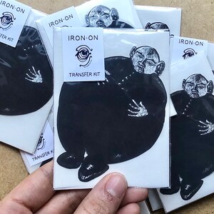 May include: A set of iron-on transfer kits featuring a cartoon character with a large head and a small body. The character is wearing a black shirt and has a white eye. The text "IRON-ON TRANSFER KIT" is printed on each sheet.