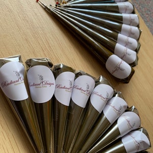May include: A close-up of several brown henna cones with white labels that say "Radiant Designs Henna Artist".