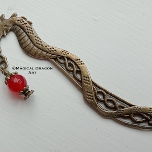 May include: A bronze dragon bookmark with a red gemstone hanging from its neck. The dragon is curled in a serpentine shape with intricate details. The bookmark is a decorative and unique gift for book lovers.