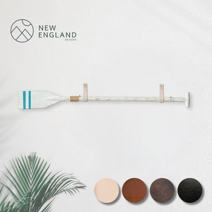 Puede incluir: Decoración de pared en forma de remo de madera blanca con rayas turquesas y un detalle de cuerda. El remo está montado en la pared con correas de cuero. El logotipo de New England Designs está en la esquina superior izquierda.