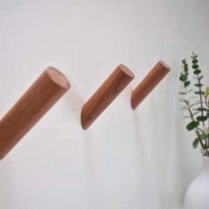 Puede incluir: Tres ganchos de pared de madera de color marrón claro, montados en una pared blanca. Los ganchos son cilíndricos y están ligeramente inclinados hacia arriba.