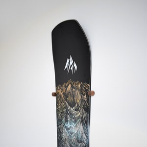 Pode incluir: Um snowboard preto com um gráfico de uma cordilheira e um logotipo branco que diz "Arbor" no topo. O snowboard está montado em uma parede com dois suportes de madeira.