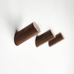 Puede incluir: Tres ganchos de pared de madera con acabado cobre. Los ganchos son de forma cilíndrica y están montados en una pared blanca.