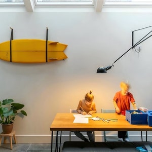 K&ouml;nnte beinhalten: Zwei Kinder sitzen an einem Holztisch mit einem gelben Surfbrett, das an der Wand hinter ihnen h&auml;ngt. Der Tisch hat ein schwarzes Metallgestell und eine blaue Aufbewahrungsbox auf der rechten Seite. Eine schwarze Lampe mit einem wei&szlig;en Schirm h&auml;ngt von der Decke.