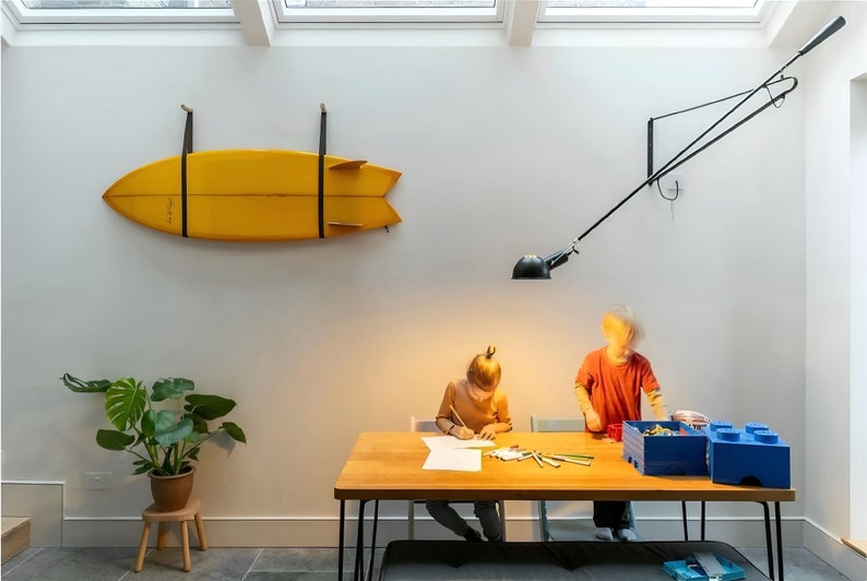 K&ouml;nnte beinhalten: Zwei Kinder sitzen an einem Holztisch mit einem gelben Surfbrett, das an der Wand hinter ihnen h&auml;ngt. Der Tisch hat ein schwarzes Metallgestell und eine blaue Aufbewahrungsbox auf der rechten Seite. Eine schwarze Lampe mit einem wei&szlig;en Schirm h&auml;ngt von der Decke.