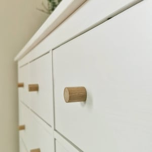 Może przedstawiać: Biała komoda z wieloma szufladami i drewnianymi gałkami. Szuflady są zamknięte, a gałki są wykonane z jasnego drewna.