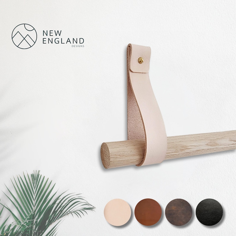 May include: A light pink leather strap with a brass screw is attached to a wooden dowel rod. The strap is looped around the rod and secured with a screw. The rod is mounted to a white wall. The text "NEW ENGLAND DESIGNS" is visible in the top left corner of the image.  Five color swatches are shown below the rod: light pink, brown, dark brown, black, and dark gray.