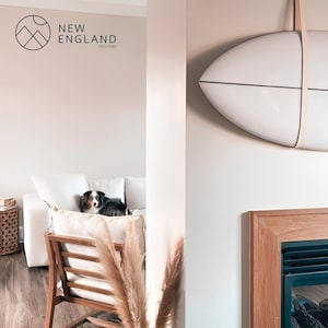 K&ouml;nnte beinhalten: Ein wei&szlig;es Surfbrett h&auml;ngt an einer Wand in einem Wohnzimmer mit einem wei&szlig;en Sofa, einem Hund und einem Kamin aus Holz. Der Text "NEW ENGLAND DESIGNS" ist an der Wand sichtbar.