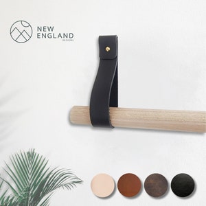 May include: A black leather towel bar holder with a brass screw, mounted on a wooden dowel. The holder is attached to a white wall.  The image also shows five color options for the leather holder: light pink, brown, dark brown, black, and dark gray.