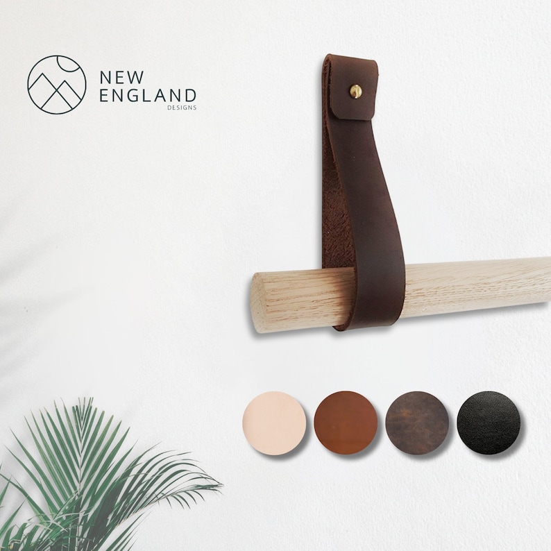 K&ouml;nnte beinhalten: Ein braunes Lederband mit einem Messingniet, befestigt an einem Holzstift. Das Band ist als Handtuchhalter oder dekorativer Wandhaken gedacht. Das Band ist in verschiedenen Farben erh&auml;ltlich, darunter Beige, Braun, Dunkelbraun und Schwarz.