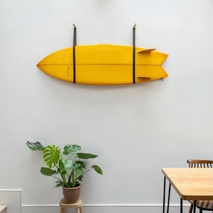 K&ouml;nnte beinhalten: Ein gelbes Surfbrett h&auml;ngt an einer wei&szlig;en Wand mit schwarzen Riemen. Eine gr&uuml;ne Pflanze in einem braunen Topf steht auf einem Holzhocker. Ein Holztisch mit schwarzen Metallbeinen steht im Vordergrund.