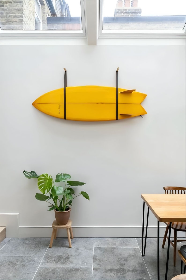 K&ouml;nnte beinhalten: Ein gelbes Surfbrett h&auml;ngt an einer wei&szlig;en Wand mit schwarzen Riemen. Eine gr&uuml;ne Pflanze in einem braunen Topf steht auf einem Holzhocker. Ein Holztisch mit schwarzen Metallbeinen steht im Vordergrund.